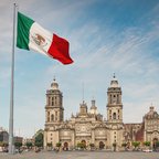 The Mexican flag waving in the foreground of the Zócalo plaza, with the historic Metropolitan Cathedral of Mexico City in the background under a blue sky.