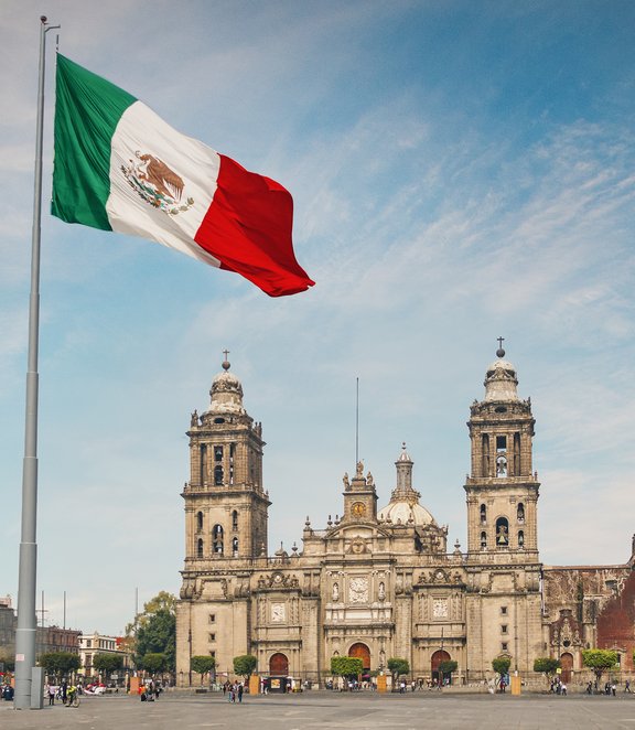 The Mexican flag waving in the foreground of the Zócalo plaza, with the historic Metropolitan Cathedral of Mexico City in the background under a blue sky.
